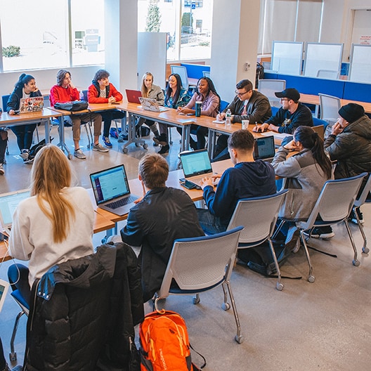 A group of students sit around a large, U-shaped table in a classroom, using laptops and notebooks while listening to a speaker. Bright daylight streams in through the windows.