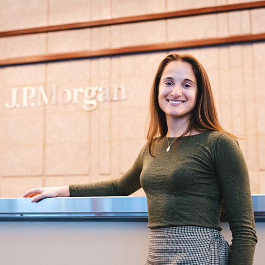 A woman with long brown hair, wearing a green top and plaid skirt, smiles while standing at a counter in front of a J.P. Morgan sign on a beige wall.