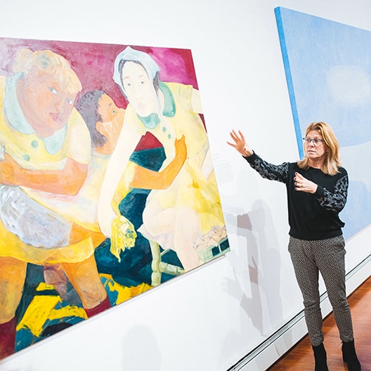 A woman stands next to a colorful painting on a gallery wall, gesturing with her hands as she speaks. The painting depicts three expressive figures in dynamic poses. Another large, blue artwork hangs on the adjacent wall.