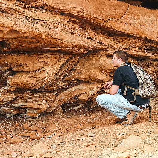 A person wearing a backpack crouches near a layered rock formation, closely examining the textured and stratified surface in a dry, rocky environment.
