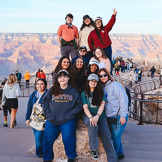 A group of twelve people pose and smile on a rocky overlook at the Grand Canyon with scenic canyon views in the background. Some are sitting, others are standing, and one person flashes a peace sign.