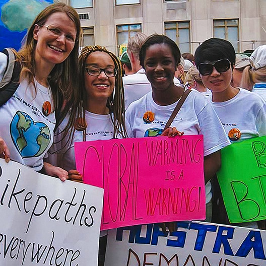 A diverse group of people at a climate protest hold colorful signs with messages like “Global Warming is a Warning!” They are smiling and standing together outdoors, wearing white T-shirts with earth graphics.