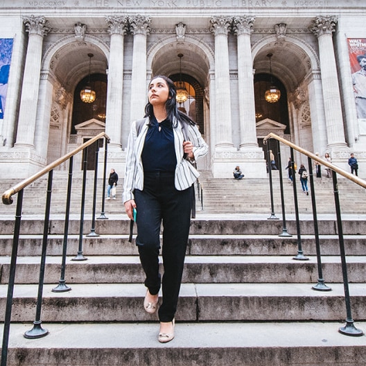 A woman in business attire walks down the steps outside the New York Public Library, with large columns and arched doorways in the background. Other people are sitting and walking near the entrance.