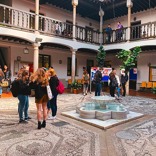 Students gather in a historic courtyard with a patterned stone floor, central fountain, and two-story colonnade. Some are talking in groups while others walk around or stand on the balcony above.