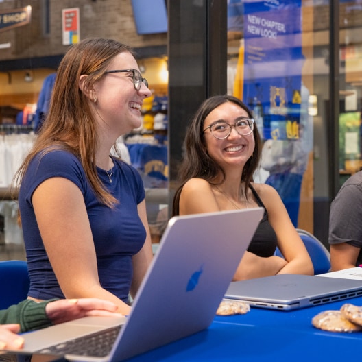 Two women sit at a table with laptops and cookies, smiling and talking. They are indoors, with a shop selling clothing in the background.