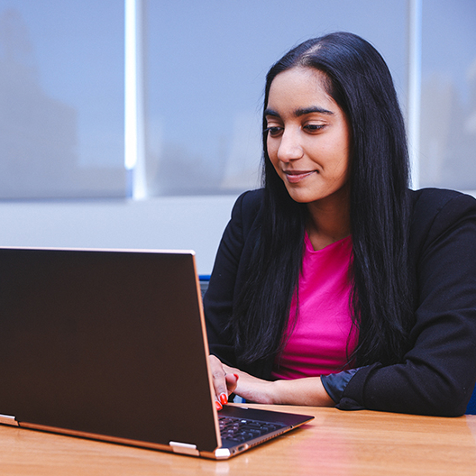 A woman with long black hair, wearing a pink top and black blazer, sits at a desk and works on a laptop in a bright, modern office space.