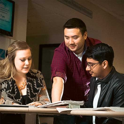 Three people sit at a table with open books and papers. One person stands, pointing at something in the book, while the other two sit and look at the material attentively. They appear to be collaborating on a project.
