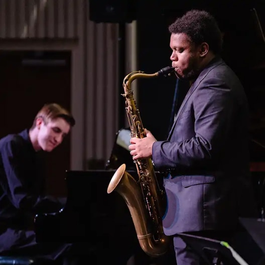 A man in a suit plays a saxophone on stage while another man in black plays the piano in the background. Both are focused on their music during a live performance.