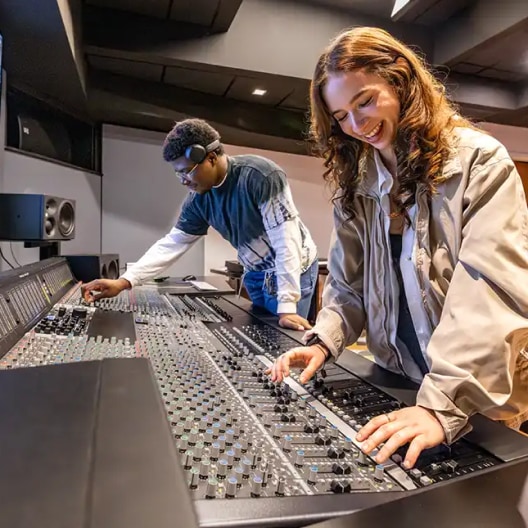 Two people in a recording studio operate a large audio mixing console, adjusting controls and smiling as they work together on sound production.