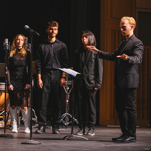 A music conductor leads three vocalists standing on stage, each dressed in black. Microphones and music stands are placed in front of them under warm stage lighting.