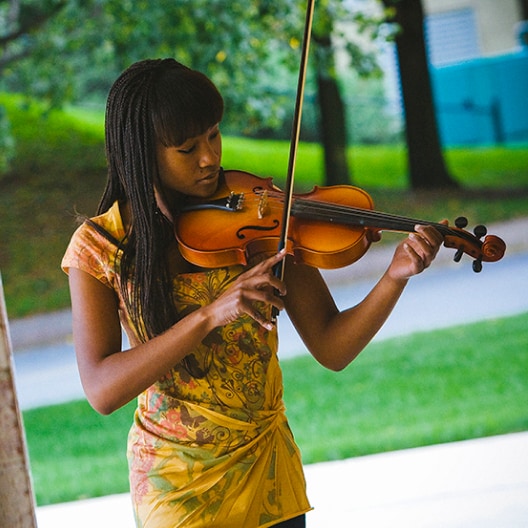 A young woman with long braids wearing a colorful dress plays the violin outdoors in a park-like setting with green grass and trees in the background.