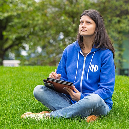 A woman in a blue hoodie sits cross-legged on grass, holding a tablet and stylus, looking up thoughtfully. Trees and greenery are in the background.