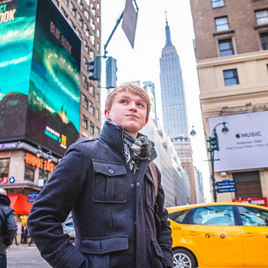 A person wearing a dark coat and scarf stands on a busy city street with yellow taxis, tall buildings, billboards, and the Empire State Building in the background.