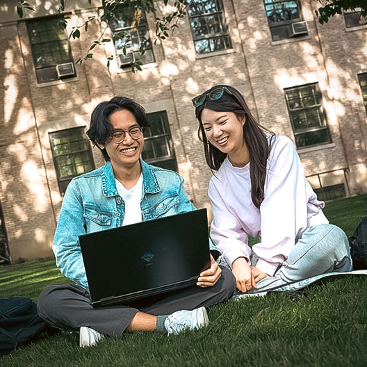 Two people sit on grass outside, smiling and looking at a laptop together. They appear relaxed and happy, with a building and tree shadows in the background.
