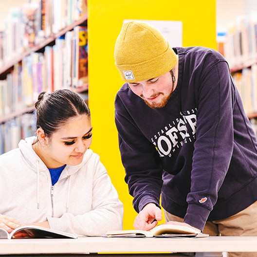 Two students in a library study together at a table; one is sitting and reading, while the other stands beside her, pointing at something in an open book. Shelves of books are visible in the background.