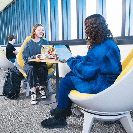 Two people sit in modern yellow chairs at a small table with laptops, smiling and talking. Another person sits in the background working on a laptop by the windows.