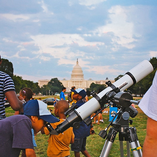 Children look through a telescope on a grassy area with the U.S. Capitol building in the background, while adults and other children stand nearby under a partly cloudy sky.