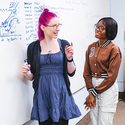 Two people stand by a whiteboard with handwritten notes. One person with pink hair is holding a marker and talking, while the other listens and smiles. Both appear engaged in discussion.