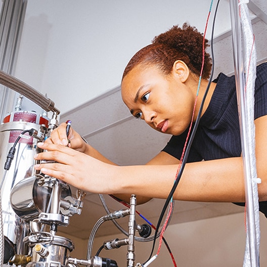 A woman adjusts wires and components on a complex scientific machine in a laboratory, focusing intently on her task.