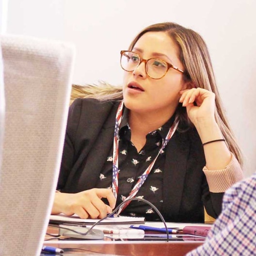 A woman with glasses and a lanyard sits at a table, speaking and gesturing with her hand. Papers and pens are on the table in front of her.