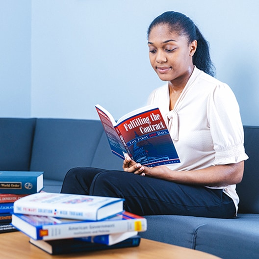 A woman sits on a blue couch reading a book titled Fulfilling the Contract. Several other books are stacked on the table in front of her. She appears focused and relaxed in a well-lit room.