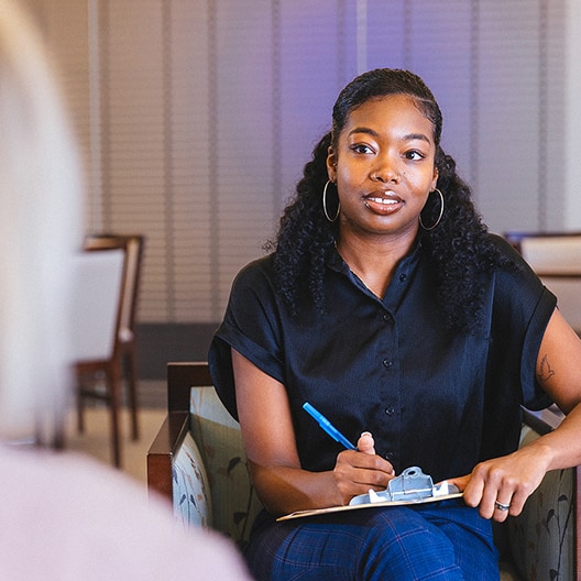 A woman with long curly hair, wearing a black shirt, sits holding a clipboard and pen, attentively listening and talking to another person in an indoor setting.