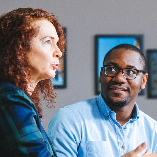 A woman with curly red hair speaks while a man in glasses and a blue shirt listens and smiles. They appear to be having a conversation indoors, with framed pictures visible in the background.