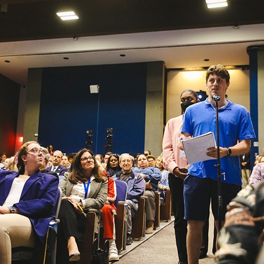 A man in a blue shirt stands with a microphone and papers, asking a question in a crowded auditorium. People seated around him listen attentively.