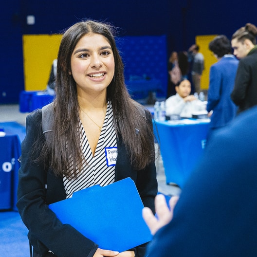 A young woman smiles while holding a blue folder at a professional event. She wears a blazer and a name tag, with people and tables in the background.