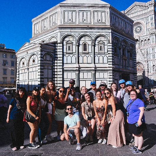 A group of people pose and smile in front of the historic, marble-paneled Florence Baptistery under a clear blue sky.