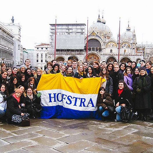 A large group of people poses outdoors holding a yellow and blue Hofstra flag, with historic buildings and domes visible in the background.