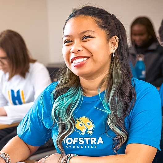 Smiling student with long, wavy, dark hair and teal highlights wears a blue Hofstra Athletics shirt, sitting in a classroom with other students blurred in the background.