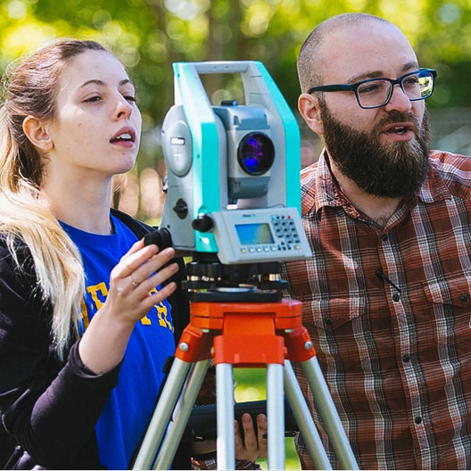 Two people outdoors use a surveying instrument on a tripod. The woman adjusts the device while the man watches. They appear focused, with green trees visible in the blurred background.