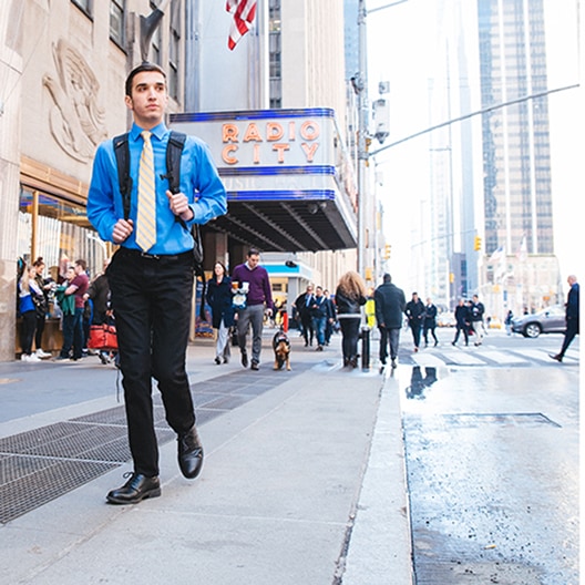 A man in a blue shirt and tie walks on a city sidewalk near Radio City Music Hall. He carries a backpack, with people and tall buildings in the background on a sunny day.