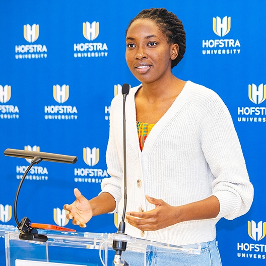 A woman speaks at a clear podium with a microphone, standing in front of a blue Hofstra University backdrop. She wears a white sweater and gestures with her hands while presenting.