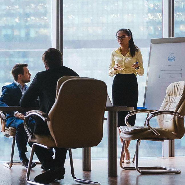 A woman stands by a flip chart, presenting to two seated men in an office with large windows and city views. The flip chart has diagrams, and the atmosphere appears professional and focused.