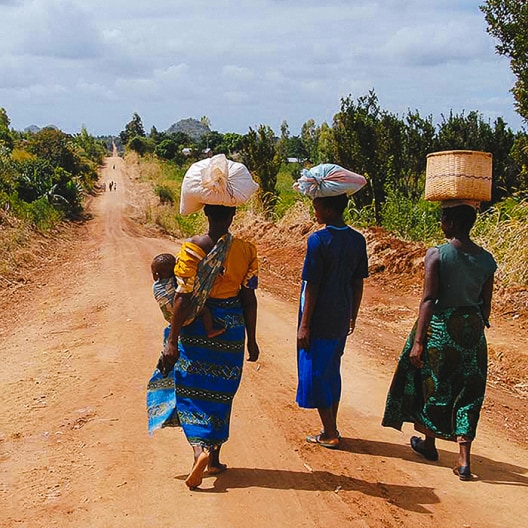 Three women walk down a rural dirt road, two with bundles on their heads and one carrying a baby on her back. Trees and bushes line the road under a cloudy sky.
