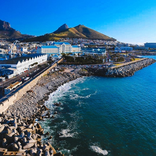 A coastal cityscape with blue water, rocky shoreline, white buildings, and mountains in the background under a clear sky.