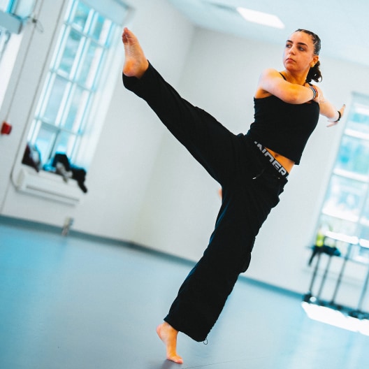 A woman in black athletic wear performs a high kick in a bright, spacious room with large windows and a smooth floor.