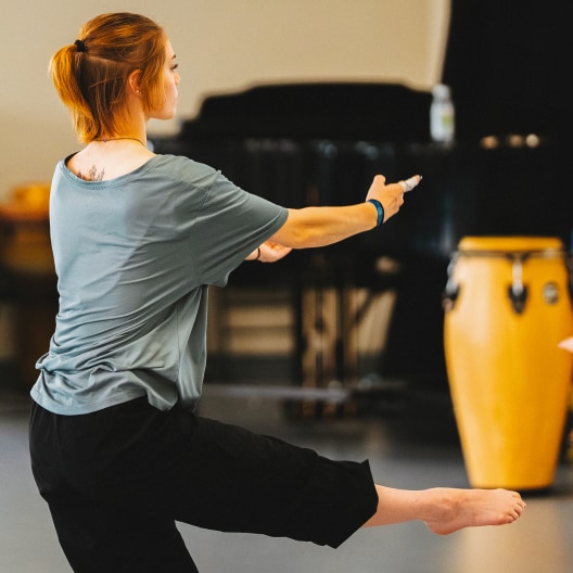 A woman with red hair in a ponytail balances on one leg with her arms extended forward in a dance studio. She is wearing a loose gray shirt and black pants. A conga drum and piano are visible in the background.