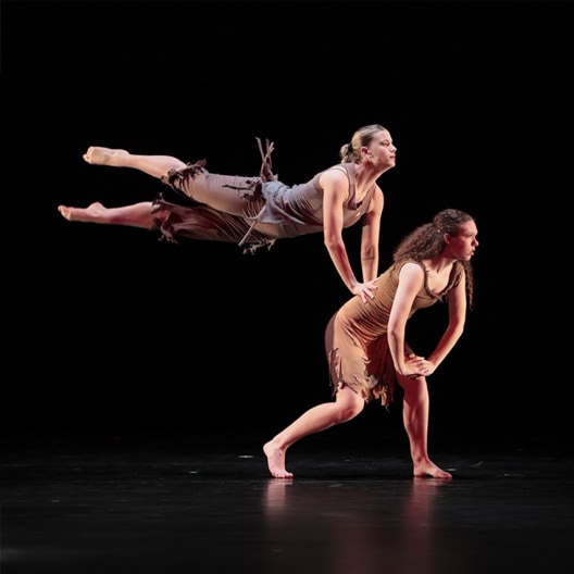 Two dancers perform on stage; one in a brown dress bends forward while supporting the other, who balances horizontally in midair with arms and legs extended, both barefoot against a dark background.