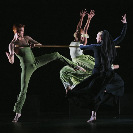 Three dancers perform on stage; one in a green bodysuit kicks while holding a staff, another in a green skirt leaps with arms raised, and a third in dark clothing moves dynamically in front of them. The background is dark.