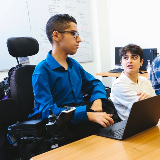 Two young men sit at a desk in a classroom, one using a wheelchair and laptop, both engaged in conversation. A whiteboard with notes and computers are visible in the background.