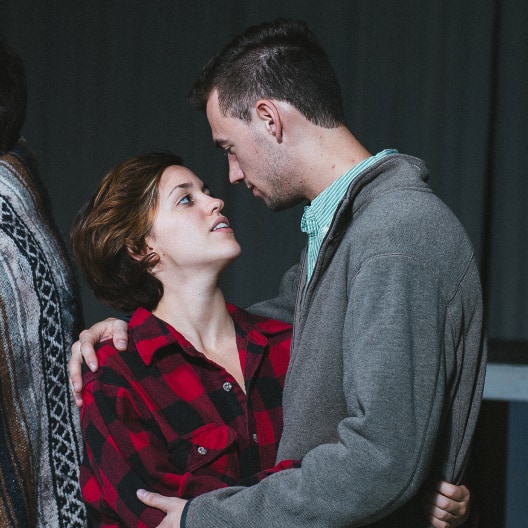 A woman in a red plaid shirt and a man in a gray jacket gaze into each others eyes, standing close together with his arm around her shoulders.