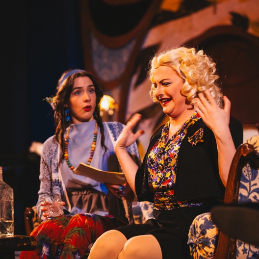 Two women sit on stage in colorful costumes. One, with long dark hair and beaded jewelry, looks surprised while holding papers. The other, with short blonde curls, gestures dramatically, smiling. The background is warmly lit.