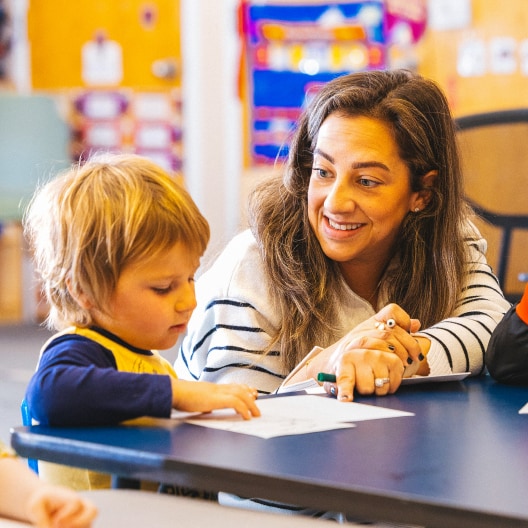 A woman smiles and sits at a table with a young child, helping him with a drawing or writing activity in a colorful classroom setting.