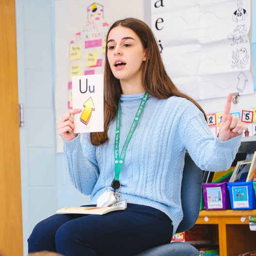 A teacher in a blue sweater sits in a classroom holding up a flashcard with the letter Uu and a picture of an up arrow, while speaking and gesturing with her hand. Classroom decorations and books are visible behind her.