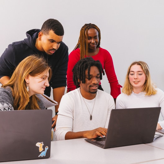 Five young adults work together around two laptops at a table, collaborating and smiling, with one person typing while others look on and engage in discussion.