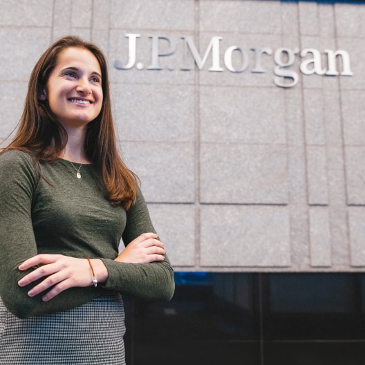 A woman with long brown hair, wearing a green sweater and gray checkered skirt, stands smiling with folded arms in front of a building wall with the sign J.P. Morgan.