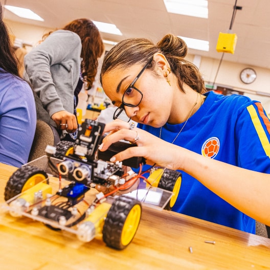 A student wearing glasses and a blue shirt works intently on assembling a small wheeled robot at a classroom table, while other students work in the background.